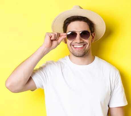 Concept of tourism and vacation. Close-up of handsome man tourist looking happy, wearing sunglasses and summer hat, standing over yellow background.