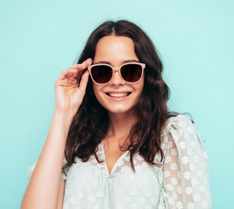 Closeup portrait of young beautiful smiling female in trendy summer dress. Sexy carefree woman posing near blue wall in studio. Positive model having fun indoors. Cheerful and happy. In sunglasses